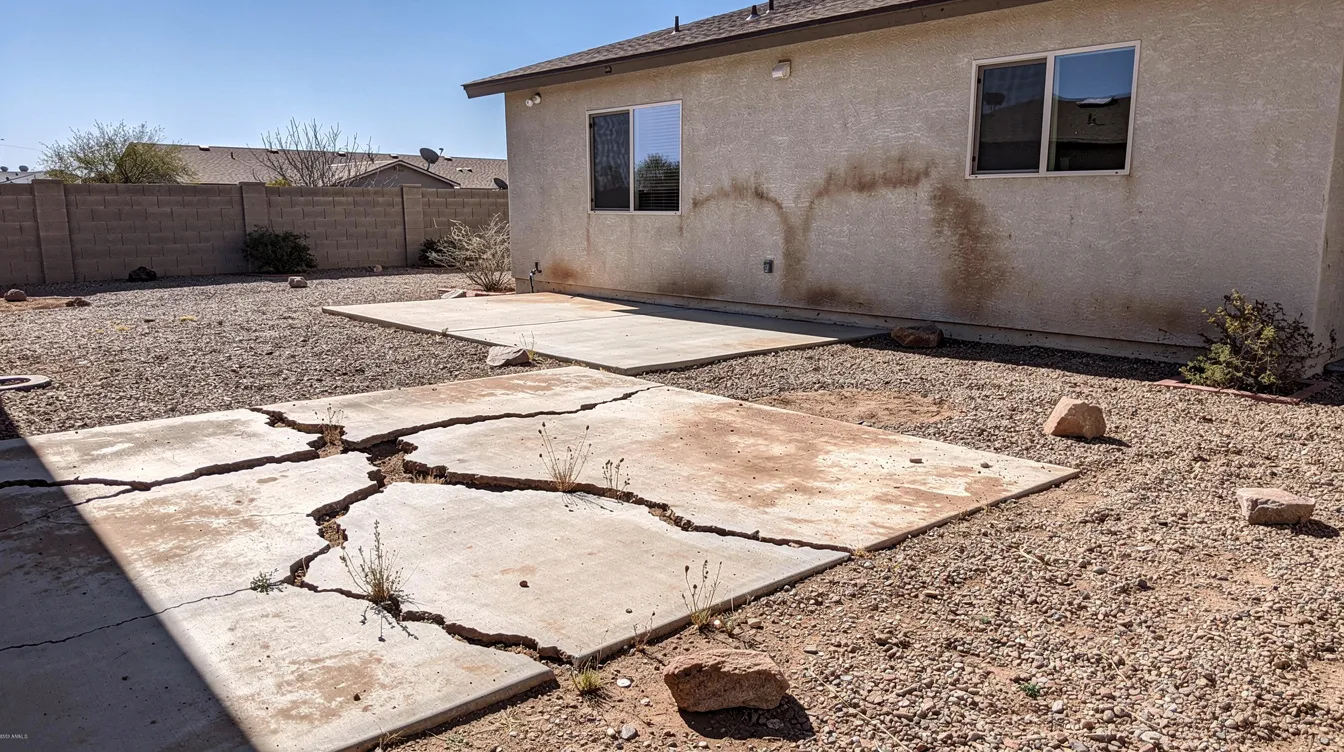 The image depicts a desert backyard in Arizona featuring cracked concrete, bare gravel, and a dusty patio area adjacent to a stucco home, illustrating a hardscaping project that emphasizes low-maintenance outdoor space. This transformed outdoor space showcases the challenges of landscaping in arid climates, highlighting the need for effective drainage solutions and quality craftsmanship.