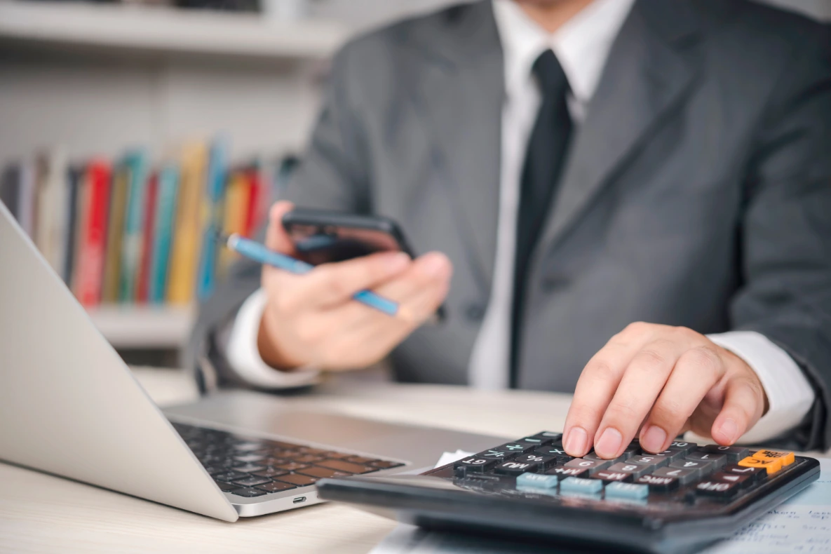 A person in a gray suit uses a calculator while holding a phone and pen, with a laptop nearby.