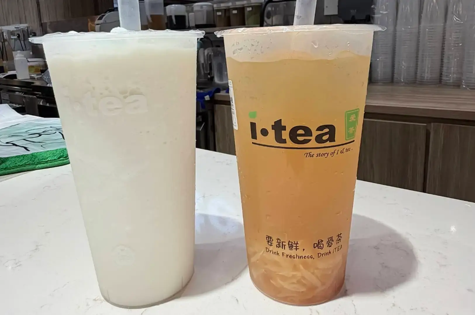 Two large iTea drinks standing on a white counter top. On the left is a tall frosted cup filled with a blended white coconut smoothie, and on the right is a clear cup displaying light orange fruit tea packed with shredded fruit pulp near the base, branded with the black "i-tea" logo and Chinese characters.
