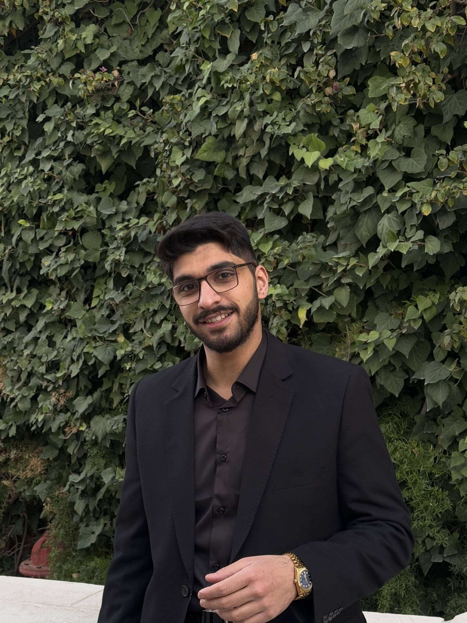 A young man with dark hair, a short beard, and glasses smiles warmly in front of a lush ivy-covered wall, dressed in an all-black suit and shirt, with a gold-tone watch on his wrist.