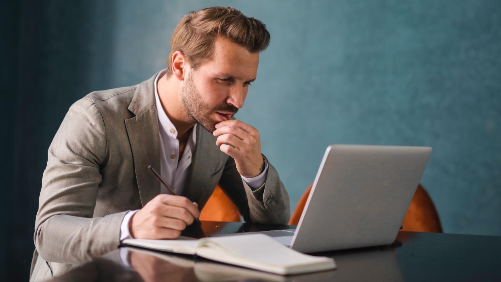 A focused business man in a gray suit works at his laptop with a pen and notebook nearby, researching FCC guidelines and wondering why he needs a POTS line replacement.