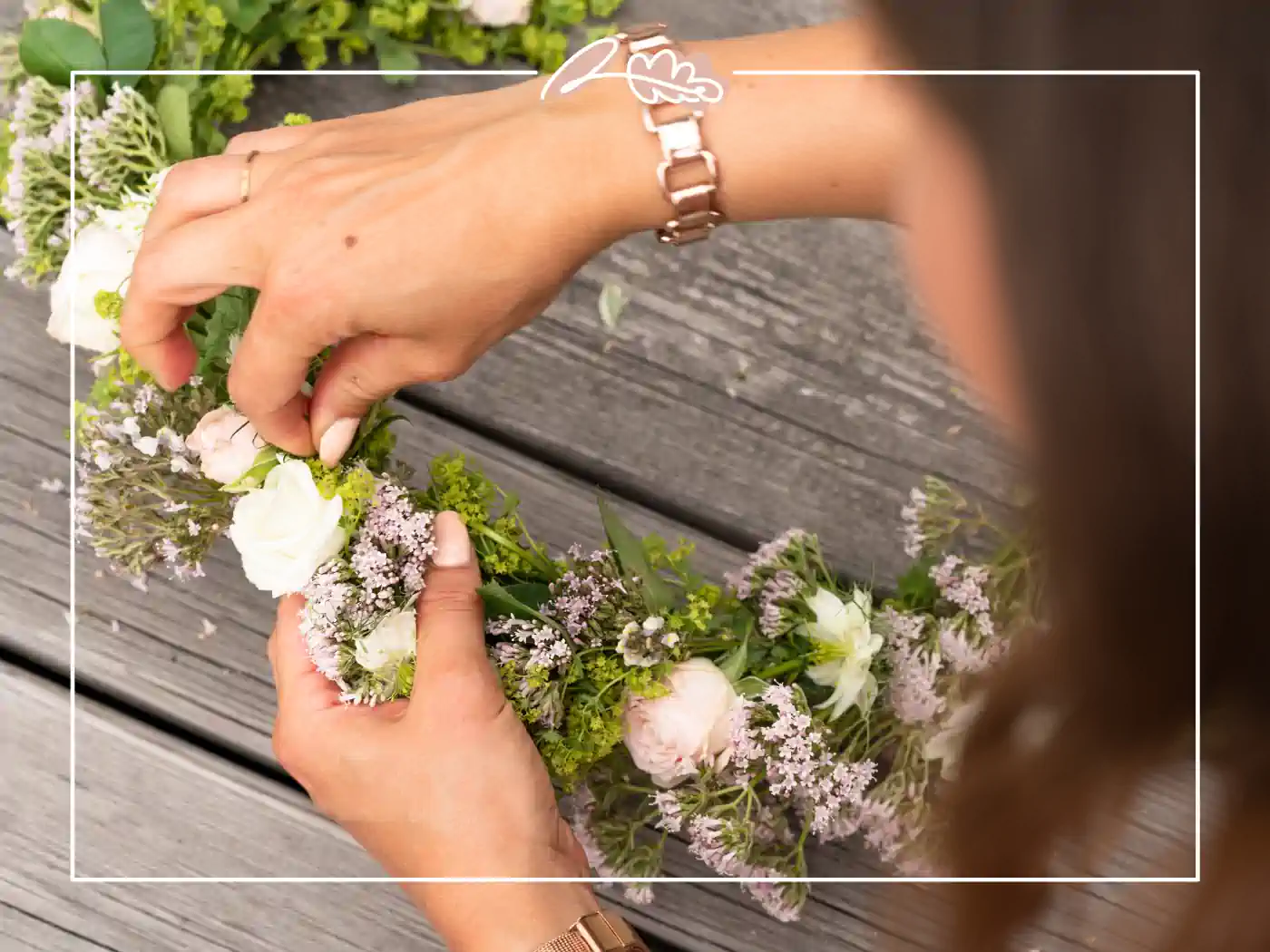 Close-up of hands delicately assembling a floral crown with white and pink blooms on a wooden surface. Blooming Romance Floral Crown by Fabulous Flowers and Gifts.