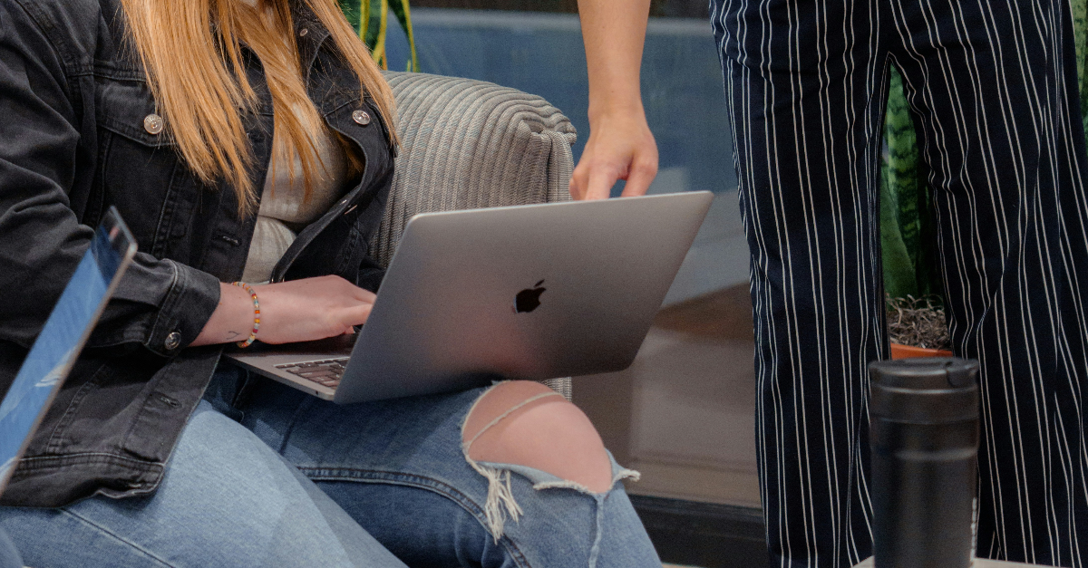 Vrouw zit met laptop op schoot, iemand staat ernaast en wijst naar het scherm