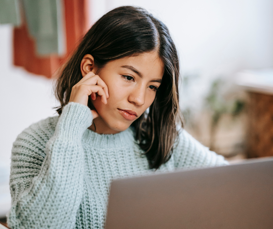 a woman in a teal sweater looks at a laptop with one hand on her face