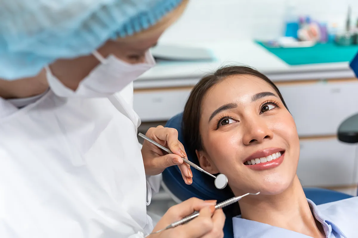 A dental professional, dentist in protective gear examining a smiling patient's teeth in a clinical setting.