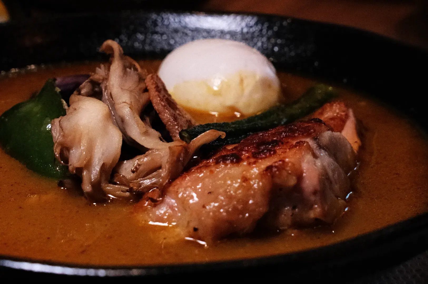 Soup curry featuring grilled chicken, sautéed mushrooms, and a poached egg in a rich sauce, served in a black skillet.