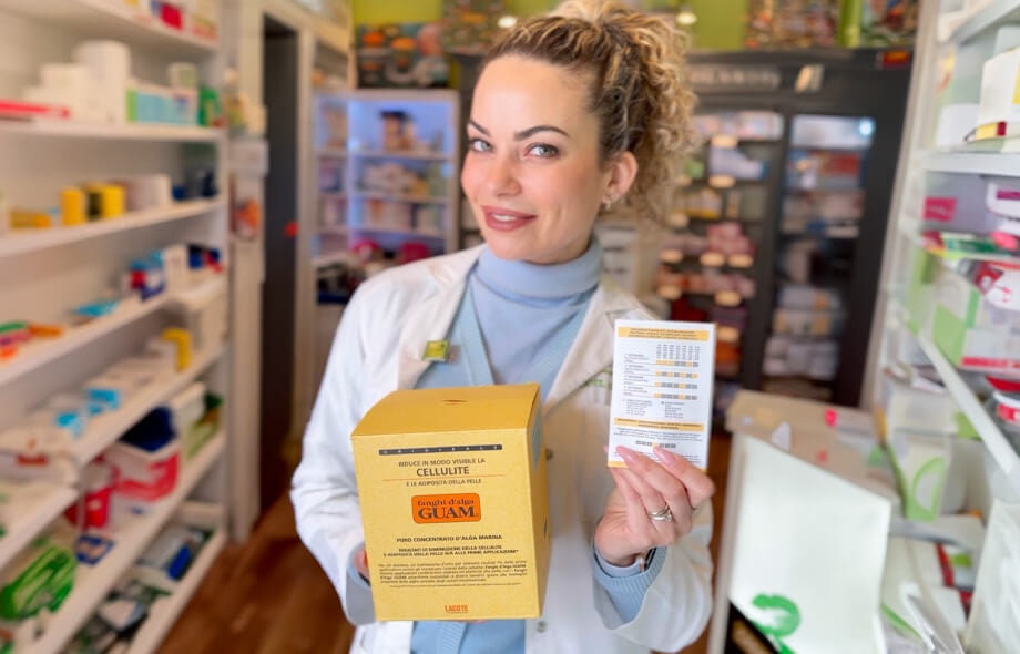 Farmacista con capelli biondi ricci raccolti che tiene nella mano destra una confezione gialla di fanghi d’alga Guam e nella mano sinistra un’altrettanta confezione con la tabella illustrativa in mostra.