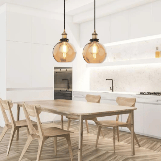 Two amber glass pendant lights hanging above a wooden dining table in a modern kitchen with white cabinets and a minimalistic design.