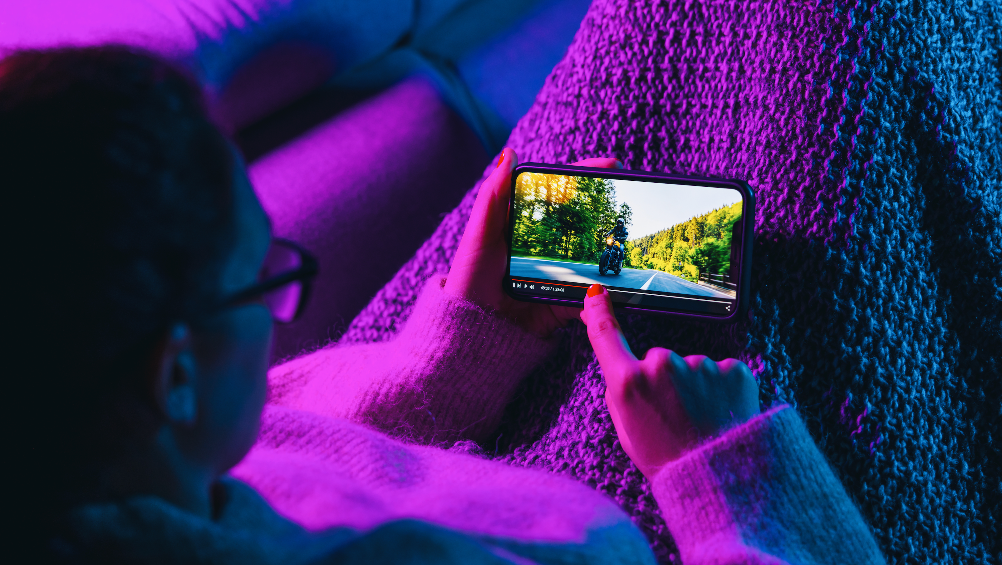 under a purple light a person with glasses streams a video on their mobile device while sitting on the couch with a blanket