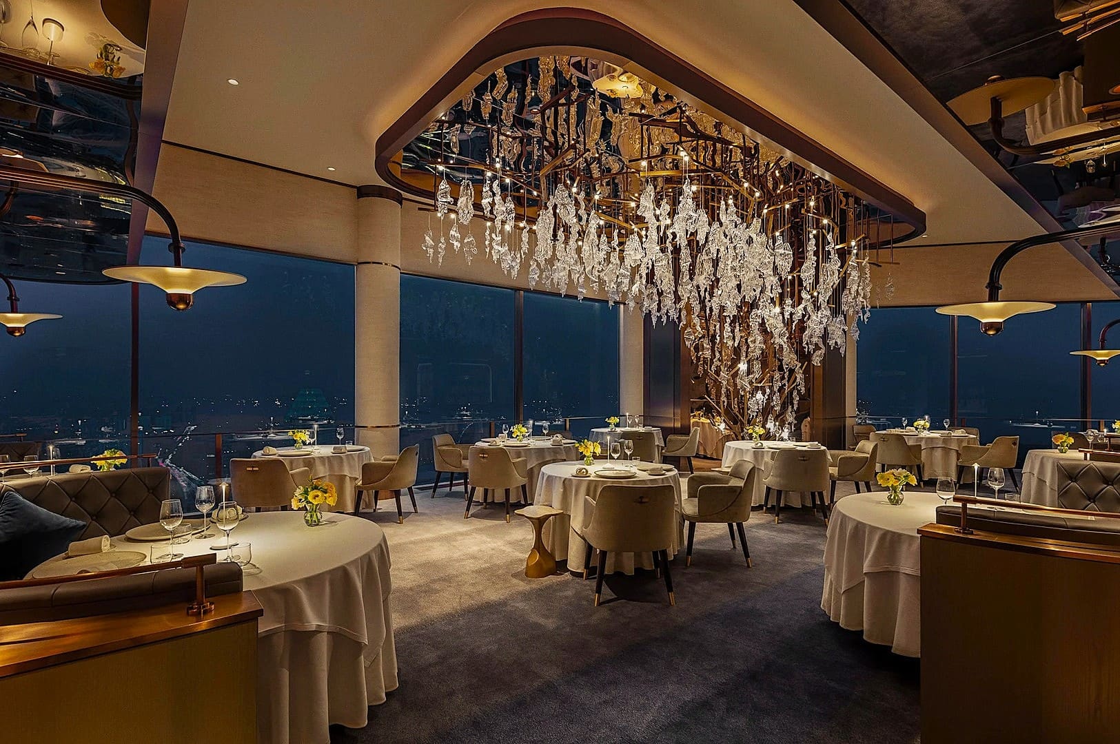 Elegant restaurant with round tables, white tablecloths, and beige chairs, under a large, artistic chandelier. Floor-to-ceiling windows reveal a city nightscape.
