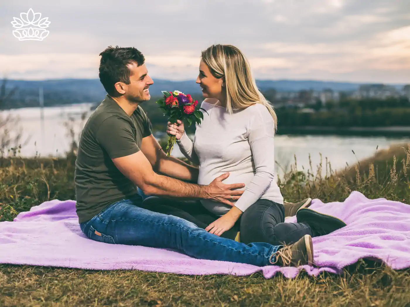 A young couple enjoying a romantic moment on a purple picnic blanket overlooking a scenic river, with the man presenting the woman a bouquet of bright flowers. Just Because Flowers. Delivered with Heart. Fabulous Flowers and Gifts.