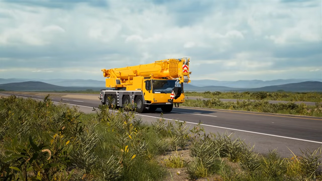 Yellow mobile crane truck driving on a highway in a scenic rural area with mountains in the background and shrubs in the foreground
