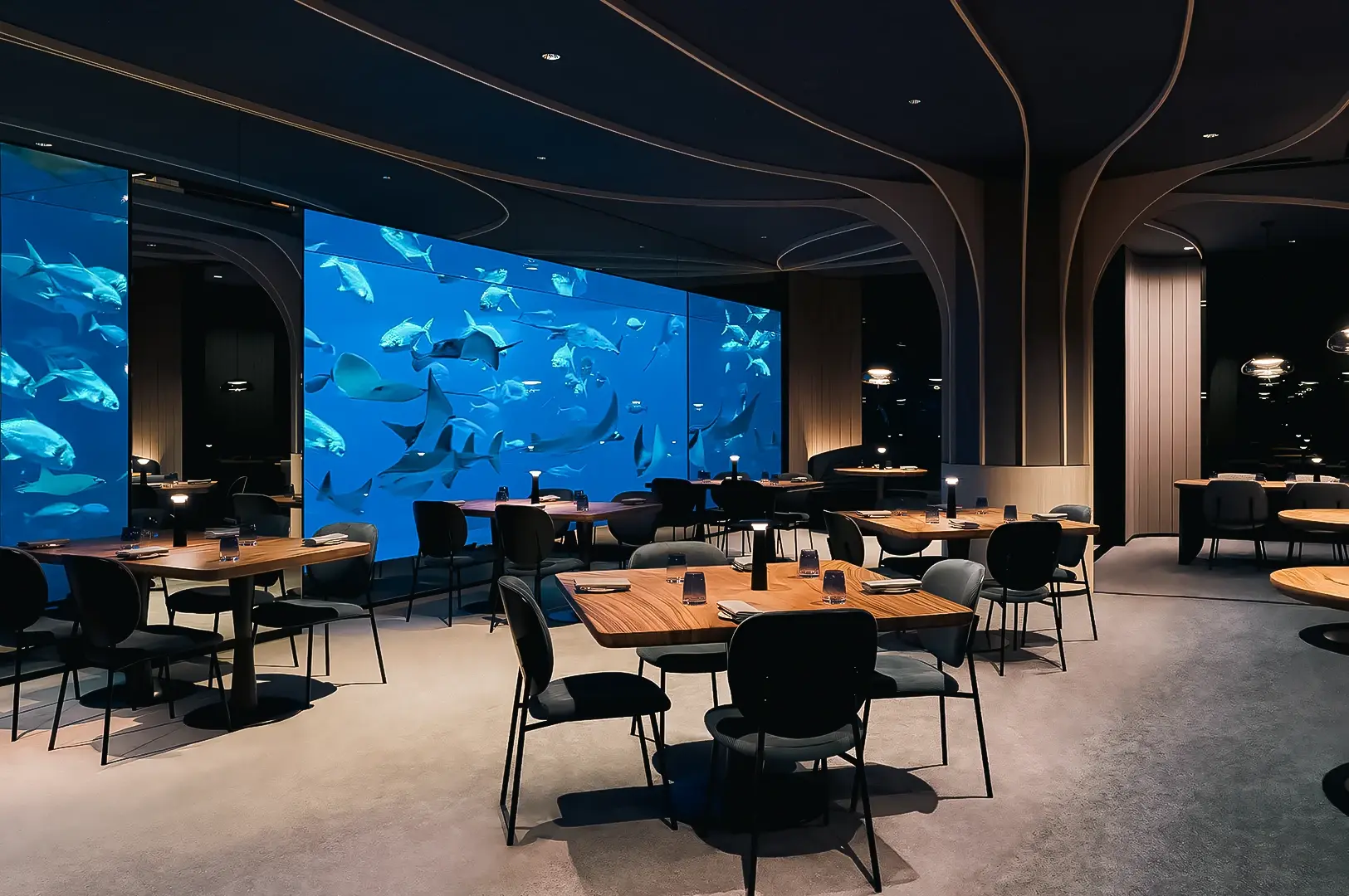 Eye‑level wide‑angle shot of a modern restaurant dining room with an immersive aquarium wall, dark architectural ceiling curves, and minimalist wooden tables and chairs.