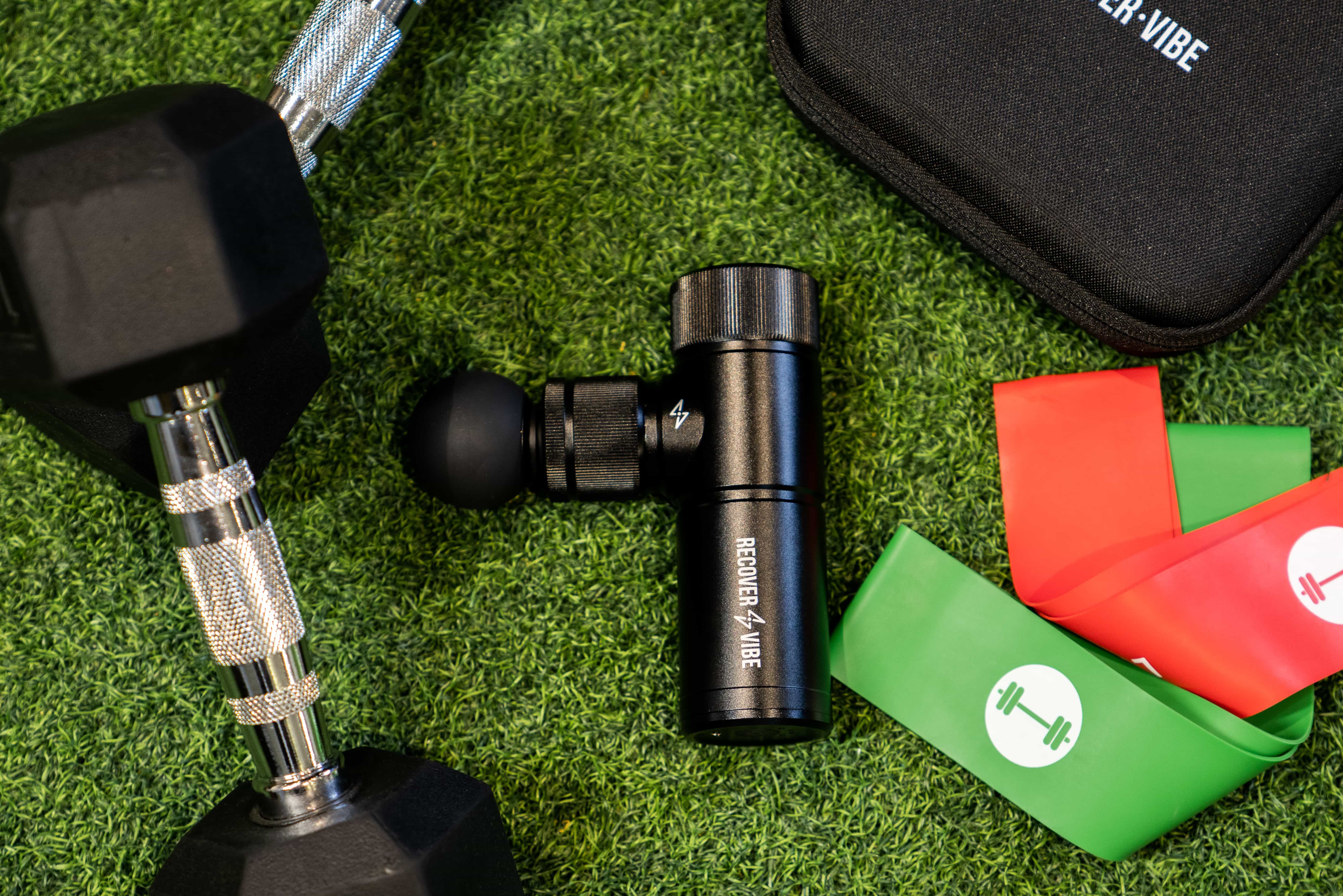 The Recover Vibe Mini massage gun on green turf next to dumbells, and elastic bands
