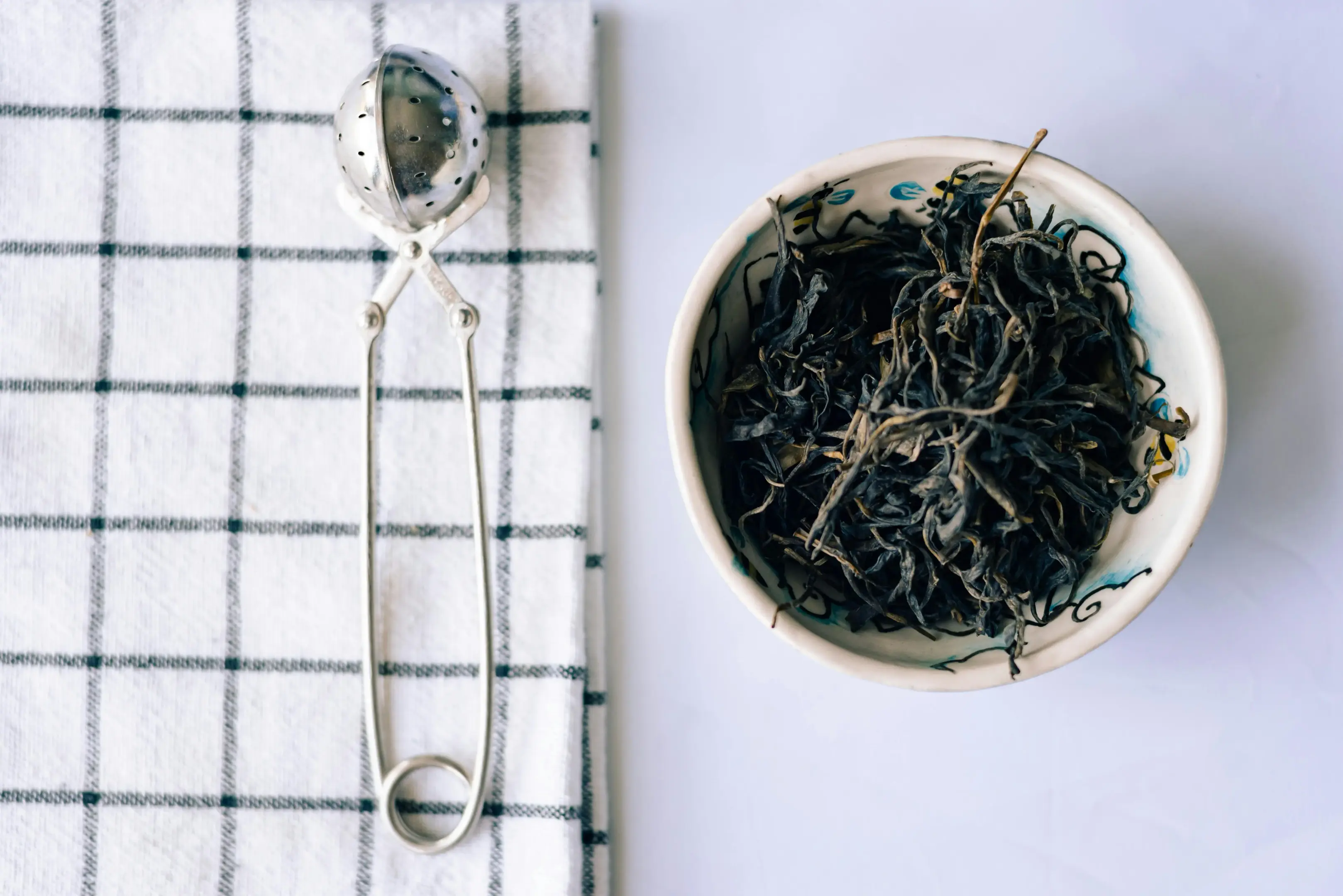 A small ceramic bowl filled with dried loose-leaf tea sits next to a stainless steel mesh tea infuser. The items are neatly arranged on a white surface alongside a blue and white checkered cloth.