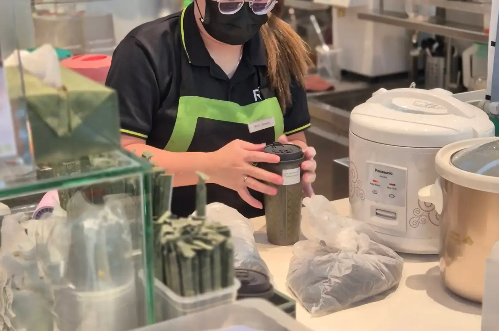 A staff member wearing a black and neon green R&B Tea uniform, face mask, and glasses secures a black lid onto a dark textured beverage cup at a clean service counter, surrounded by a Panasonic rice cooker, slow cookers, and packaged ingredients.