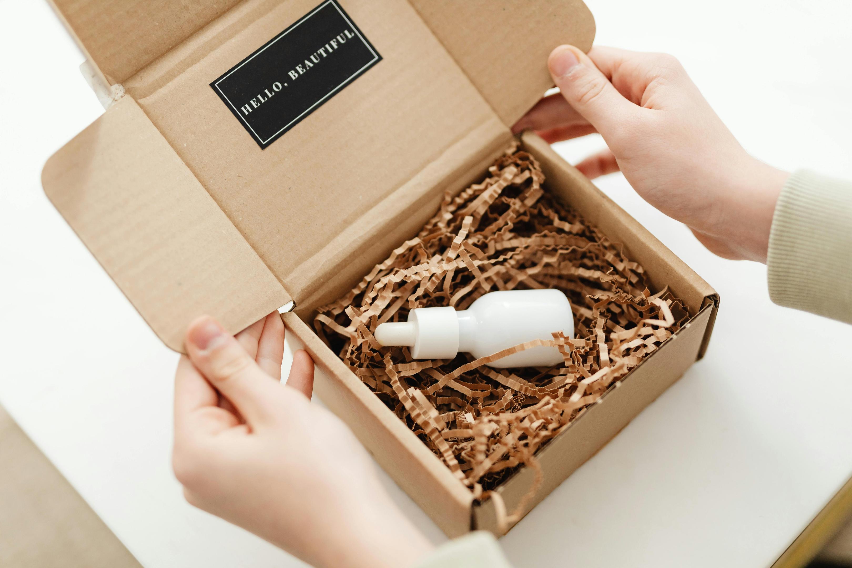 A brown gift box reveals a small white vial inside, the tag on the box reads "Hello, Beautiful."