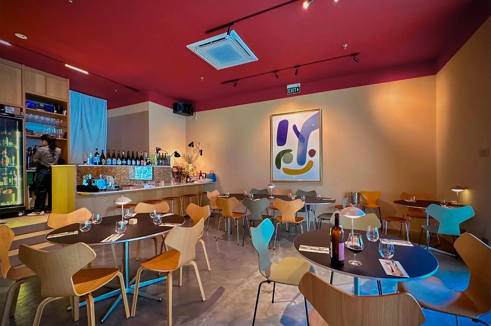 Modern cafe with wooden chairs and dark tables set for dining, featuring abstract art on a beige wall. Warm, cozy ambiance with a red ceiling.