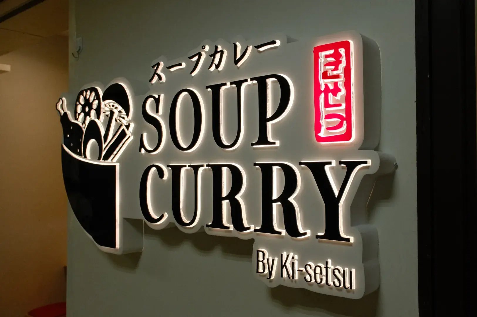 A illuminated sign outside the shop for "Soup Curry" by Ki-setsu featuring the text in Japanese on the top, and English on the bottom with a half bowl of soup curry with ingredients and utensils on the left..