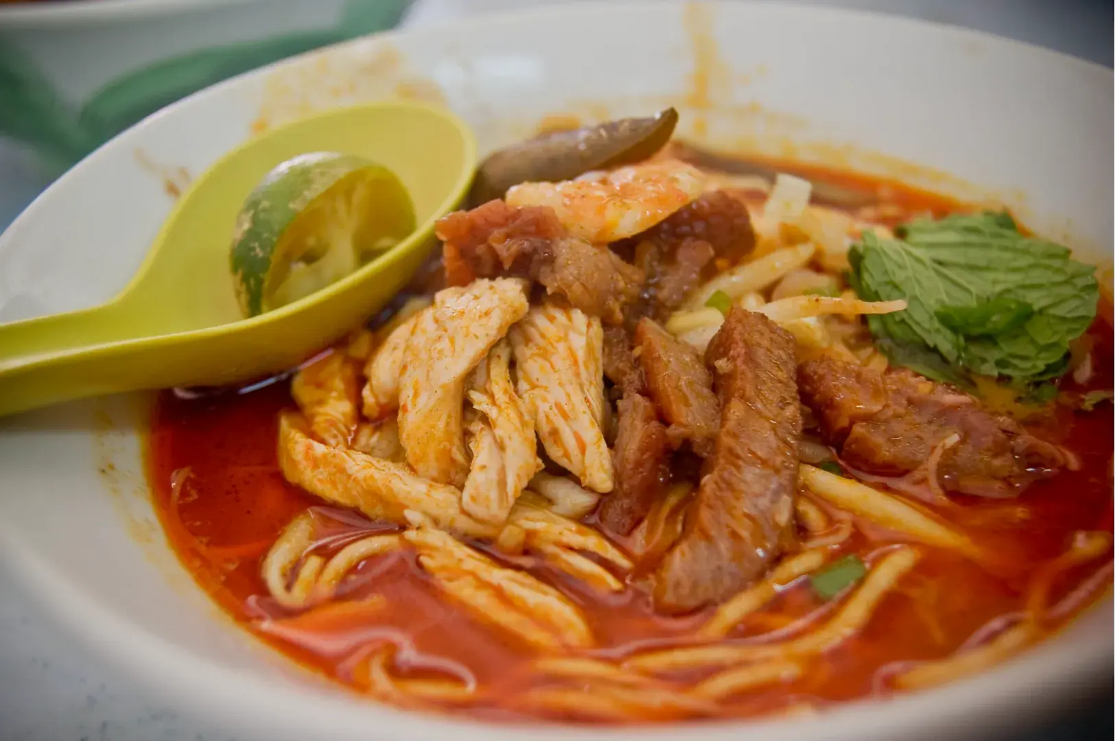 A bowl of noodles topped with sliced chicken, pork, and herbs, served in a reddish broth with a lime wedge on the side.