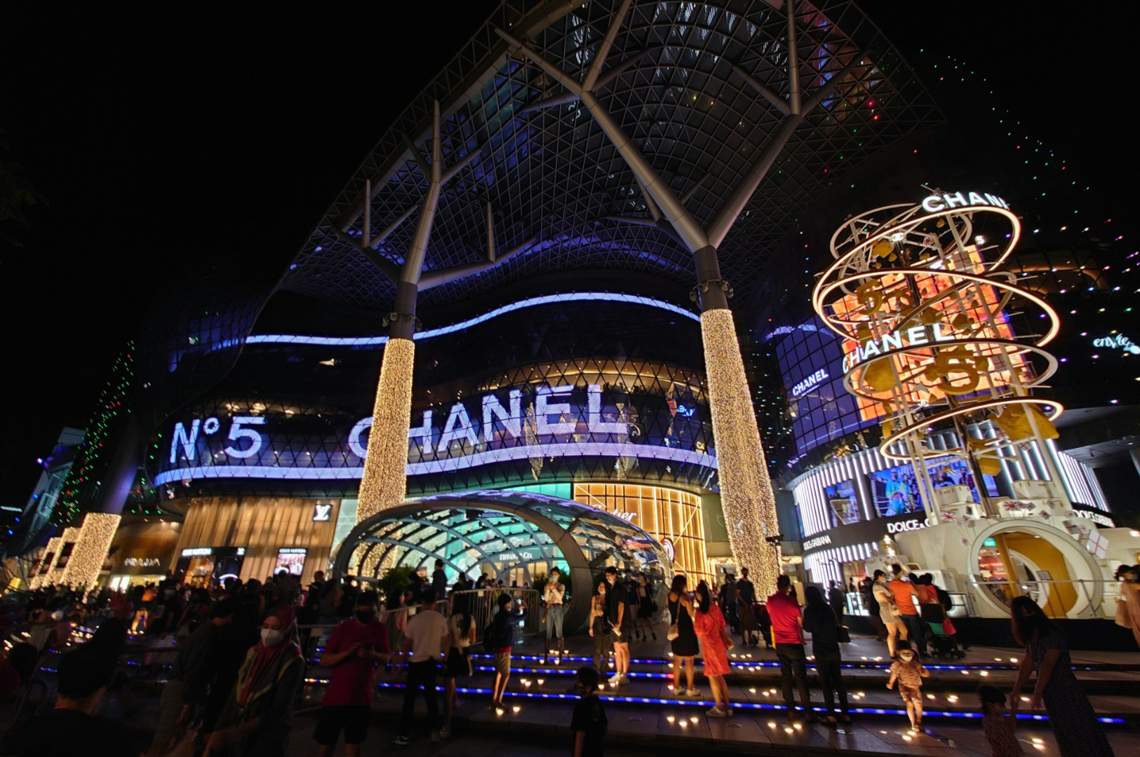 A bustling nighttime city scene featuring a modern shopping complex. Large illuminated signs read "Chanel" and "No. 5," with people gathered outside.
