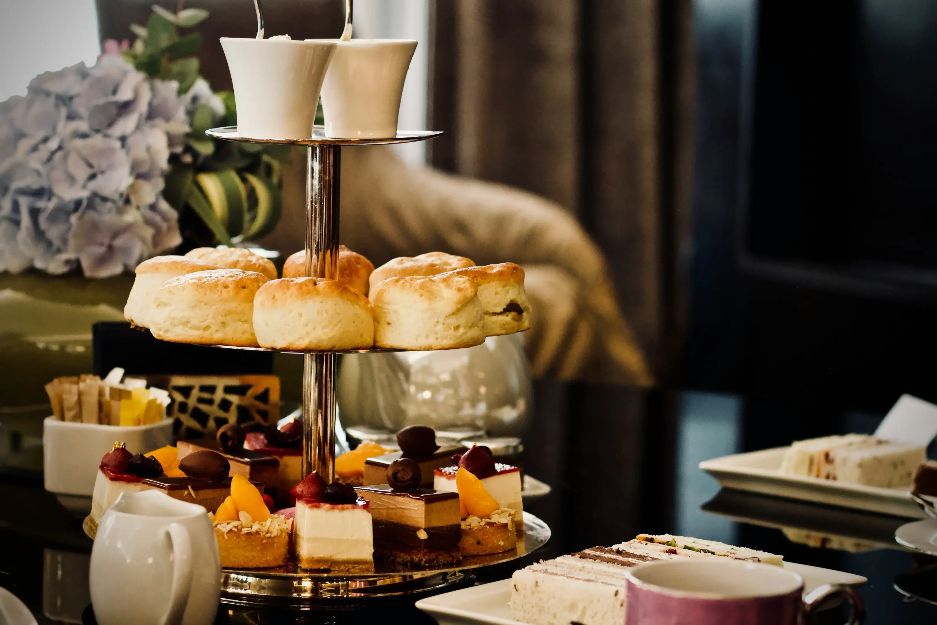 A three-tiered silver stand displays an elegant afternoon tea service featuring golden-brown scones, assorted miniature cakes, and fruit-topped pastries. Finger sandwiches and a cup of tea are arranged on the table alongside a soft blue hydrangea bouquet in the blurred background.