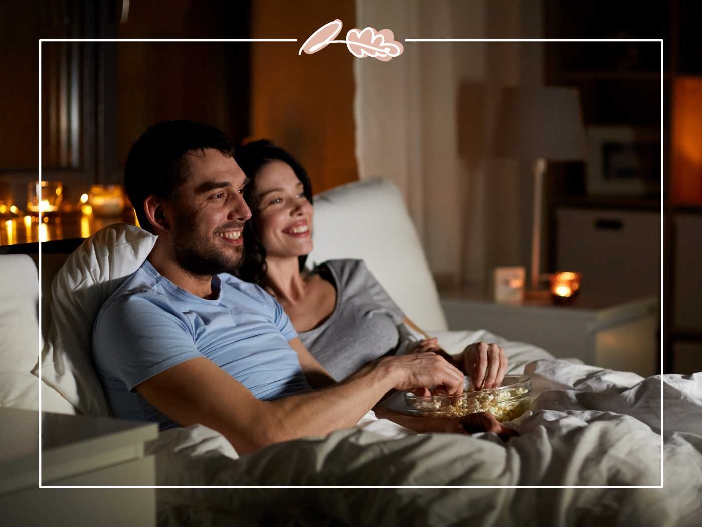 A couple in bed, smiling and enjoying popcorn while watching TV in a warm, candle-lit bedroom - Fabulous Flowers and Gifts.