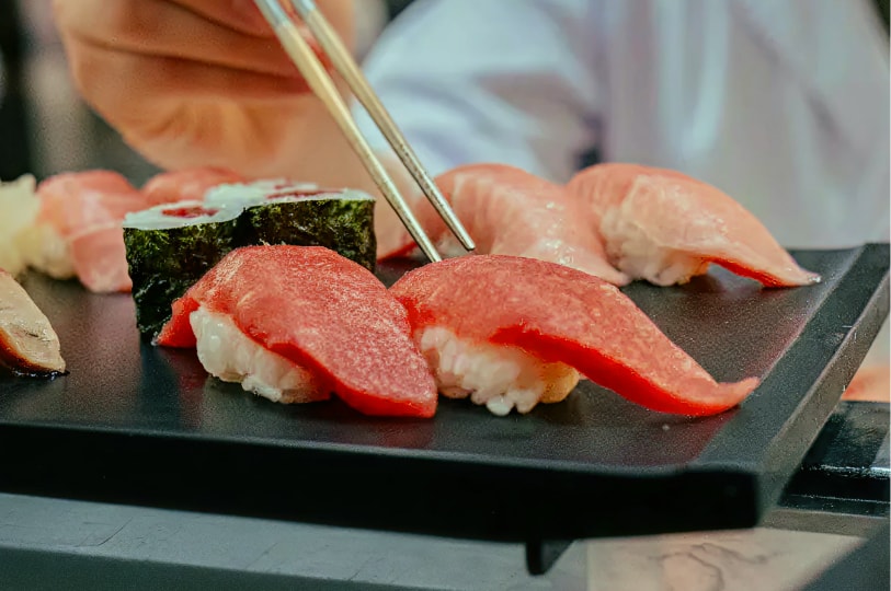 A close-up of a chef using metal plating chopsticks to arrange fresh tuna nigiri and maki rolls on a sleek black platter.