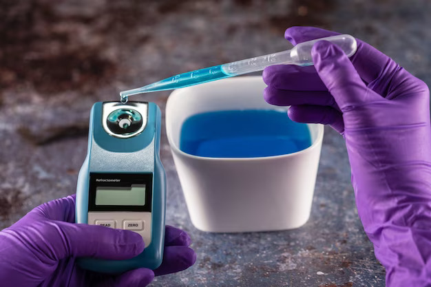 Två händer med handskar som häller blå vätska med en pipett i en digital refraktometer