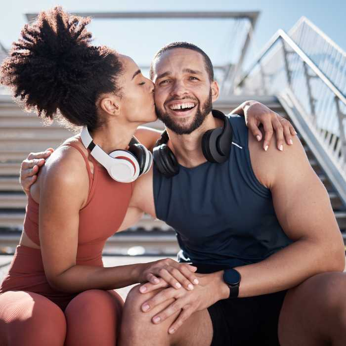 A fit couple sharing a joyful moment during a workout break on stadium steps, with the woman kissing the man on the cheek. They embody the vitality supported by the AIM Companies Supplements Collection, including options like Herbal Fiberblend and Irish moss, which promote digestive health and manage issues like constipation. Their energetic lifestyle is enhanced by products that are mindful of allergies. The Good Stuff Health Shop South Africa.