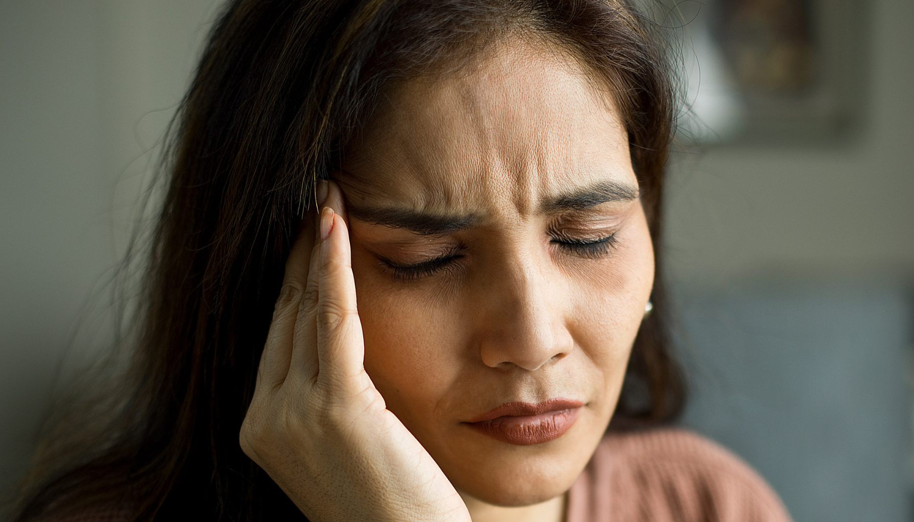 Kvinne med lukkede øyne presser hånden mot tinningen og viser tydelig tegn på hodepine eller migrene.