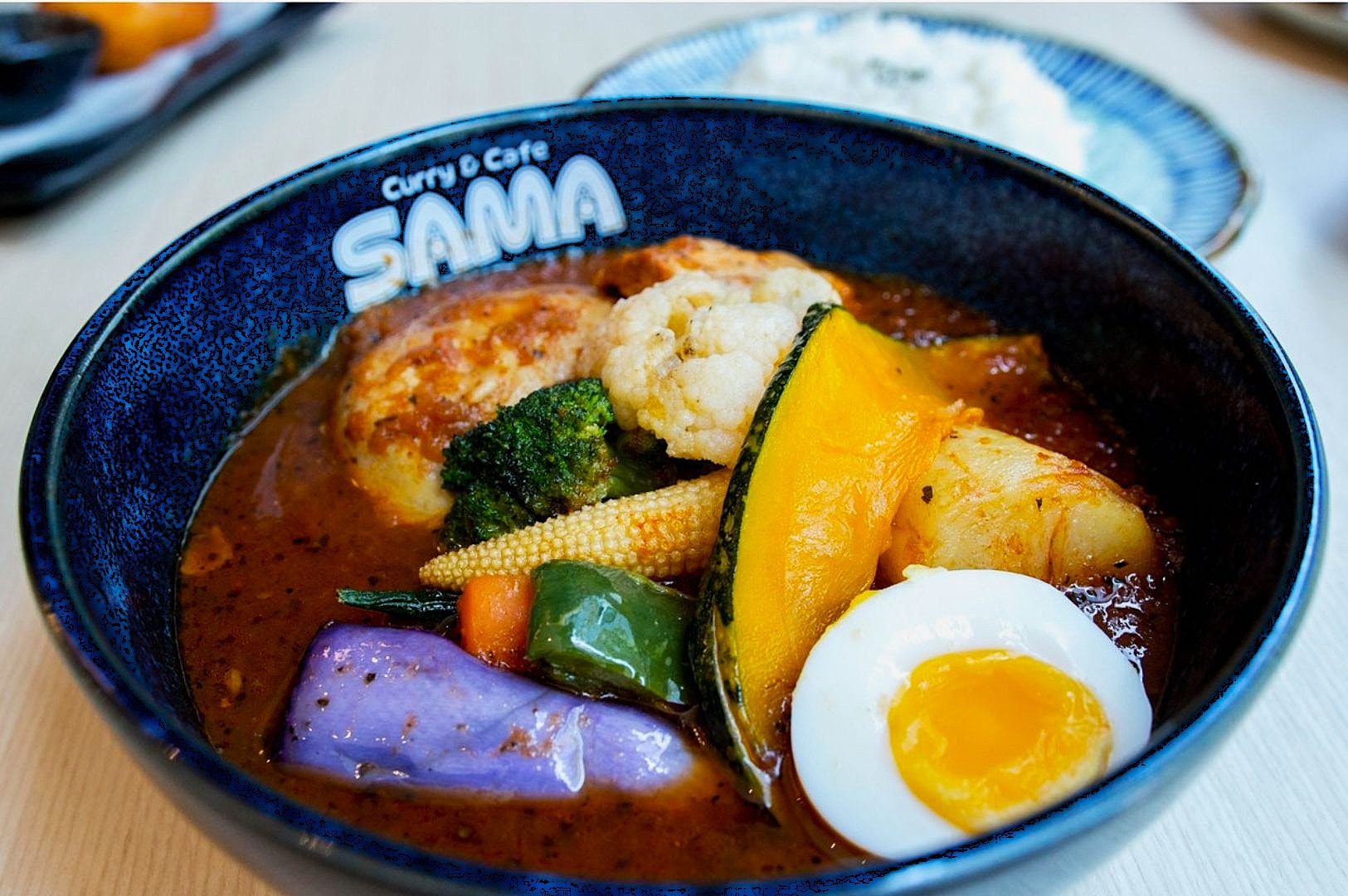 A colorful bowl of Japanese curry with vibrant vegetables like pumpkin, broccoli, eggplant, and a soft-boiled egg sits on white rice. The dish looks hearty and comforting, set in a dark blue bowl.