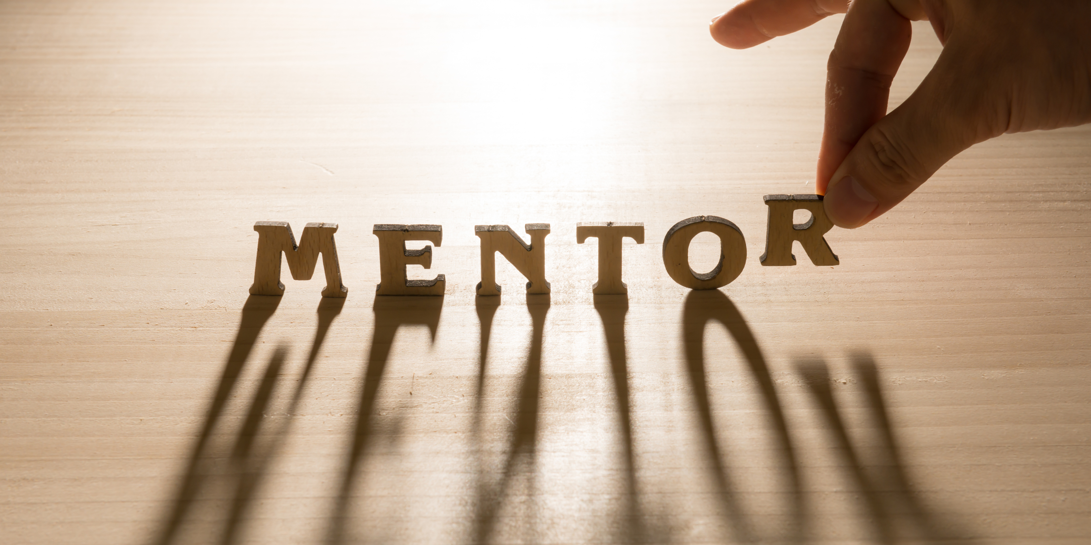 The word Mentor is placed on a wooden table with wooden letters and a hand places the last letter - R - on the table. This is being used as the header to a mentoring blog on The Marketing Mentor website.