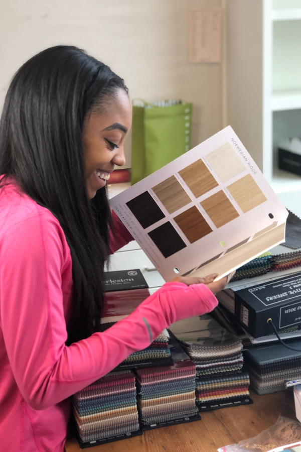 A designer in a bright pink top smiles while flipping through a wood-finish sample book, surrounded by stacks of fabric swatches.