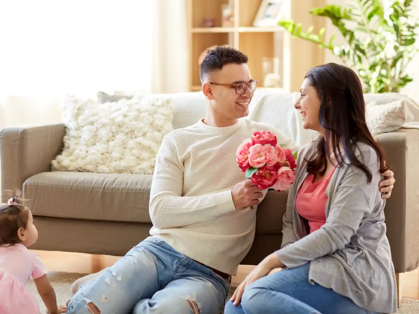 Man giving a bouquet of pink peonies to his delighted partner in their cosy living room, with their toddler watching, representing value with flower bouquets under R500, delivered with care by Fabulous Flowers and Gifts.