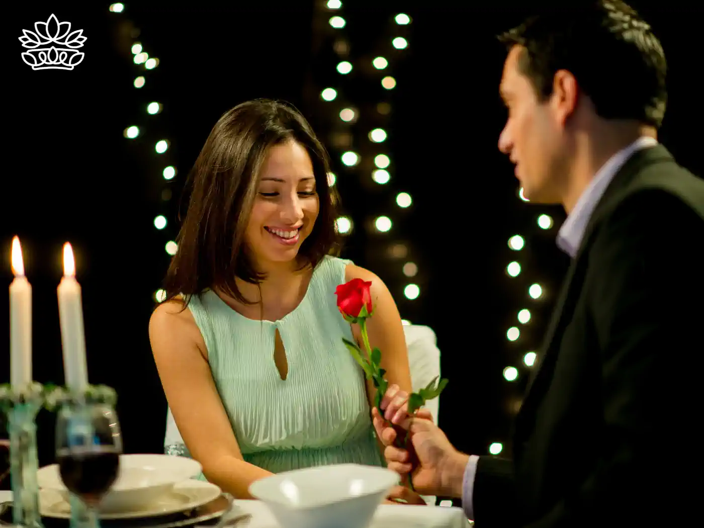 A woman smiles as she receives a romantic red rose from a man during a candlelit dinner, with twinkling lights in the background. Fabulous Flowers and Gifts delivered with heart. Expect Tips for Valentine's Day Flowers.