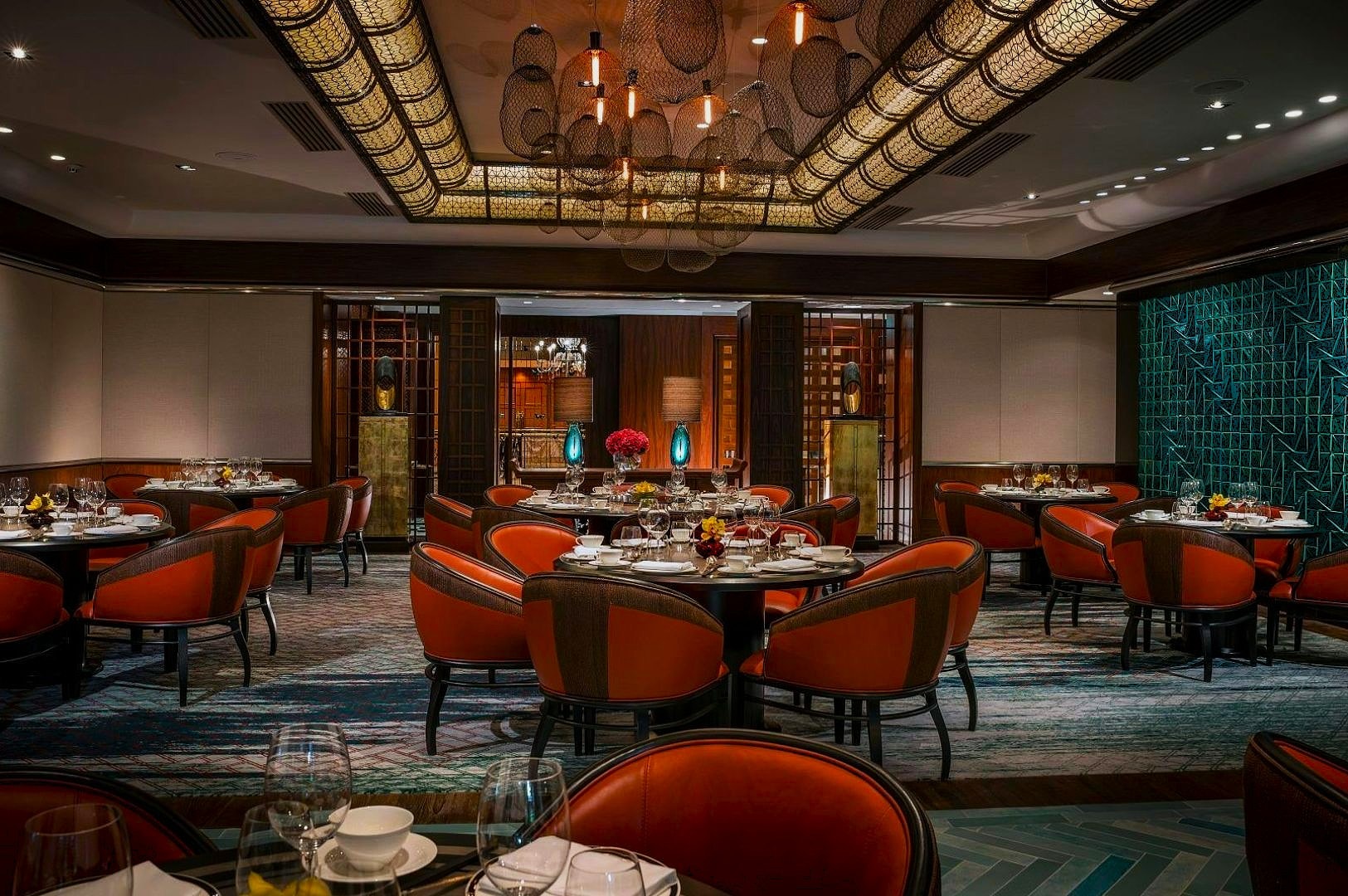 Sophisticated restaurant interior with elegant orange chairs, round tables set with glassware, and a decorative ceiling. Dim lighting creates a warm ambiance.