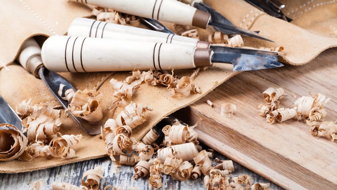 Close-up of wood carving tools with ivory-colored handles resting on a leather cloth, surrounded by wood shavings and a partially carved wooden board.
