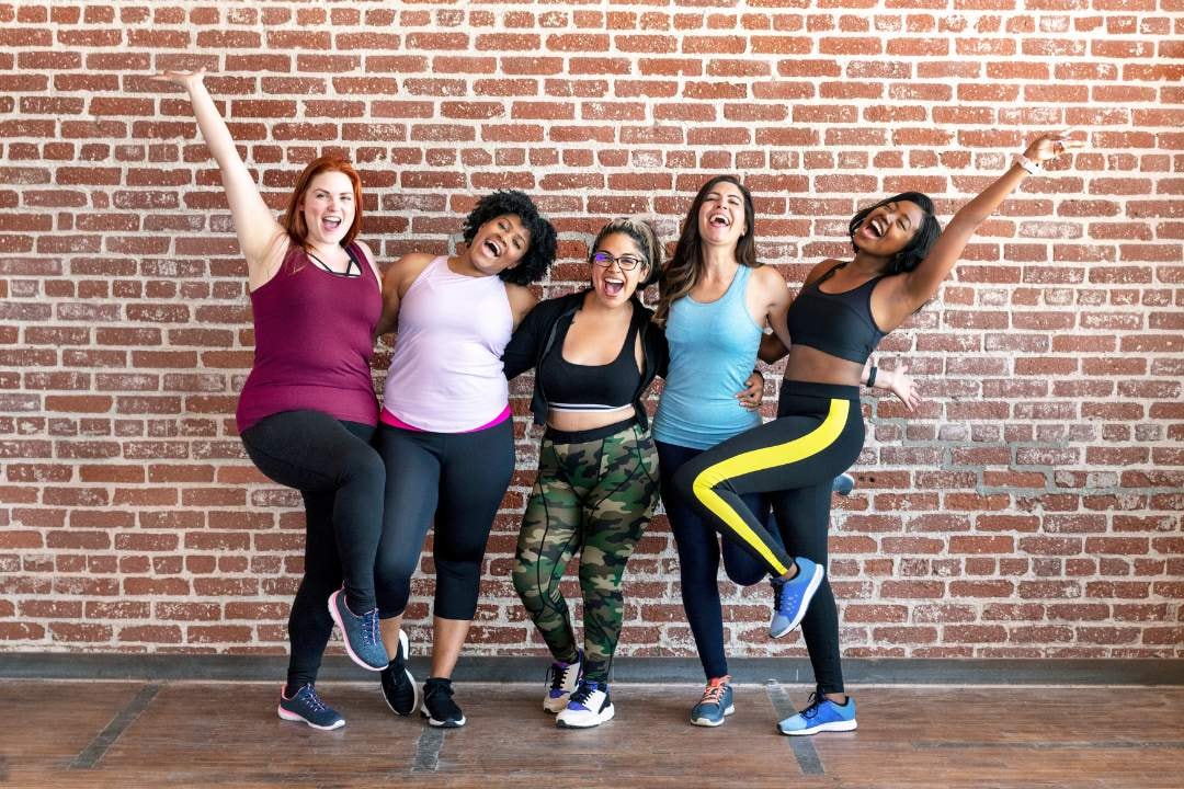 Five women standing against a brick wall, smiling and kicking their legs up in playful celebration.