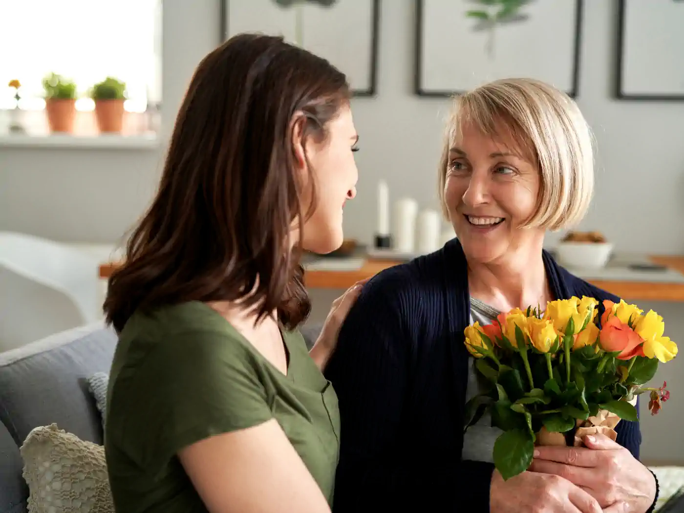 Older woman with a joyful expression receiving a vibrant bouquet of yellow and orange roses from a younger woman in a cozy home setting, sharing a moment of gratitude. Fabulous Flowers and Gifts - Thank You Flowers. Delivered with Heart.