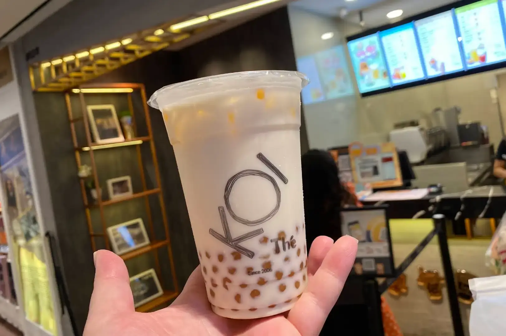 A hand holds a clear plastic cup of KOI Thé milk tea filled with ice and a generous layer of KOI's signature golden tapioca pearls resting at the bottom, set against the softly blurred background of a KOI shop interior and order counter.