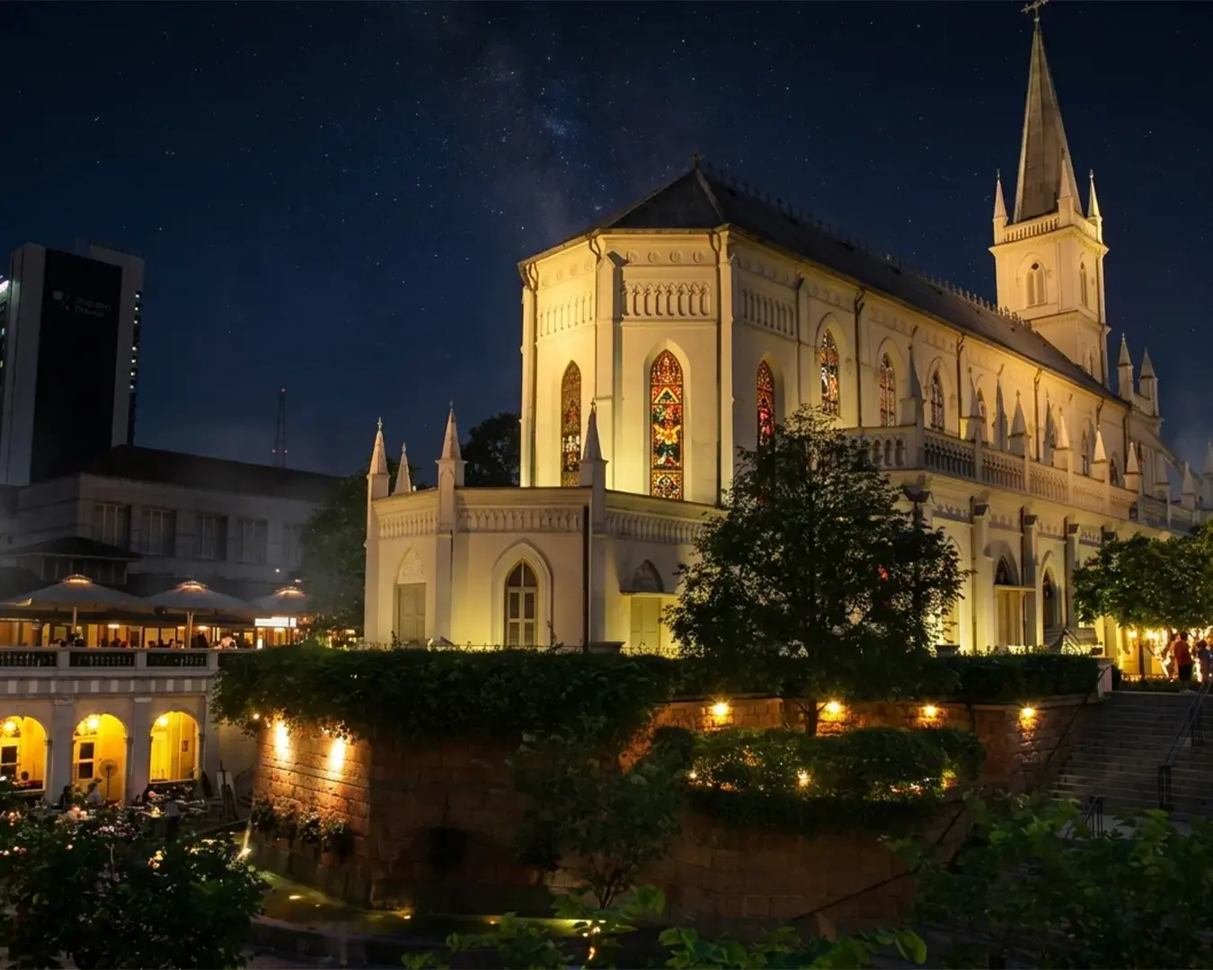 Gothic-style church illuminated at night, featuring stained-glass windows. Nearby, a lit terrace full of diners creates a lively but serene atmosphere.