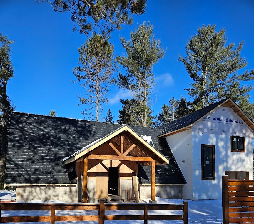 A house is enclosed by a wooden fence, with a wooden gate providing access to the property. A dark, sloped metal roof is the feature point of the house.
