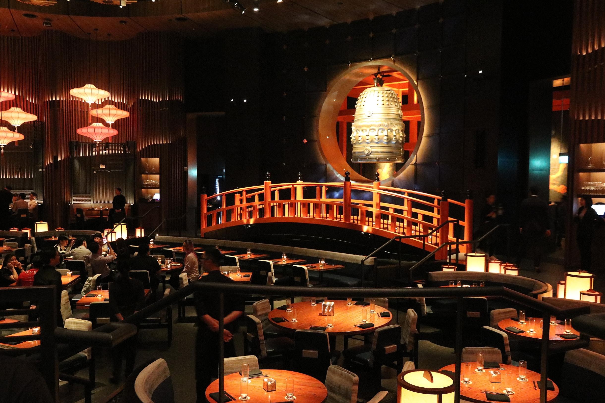  This expansive restaurant interior features a dramatic red arched bridge and a massive golden bell set within a circular wall alcove as central focal points. The dimly lit dining room is filled with patrons seated at round wooden tables, complemented by warm glow from traditional-style lanterns and tiered pendant lights.