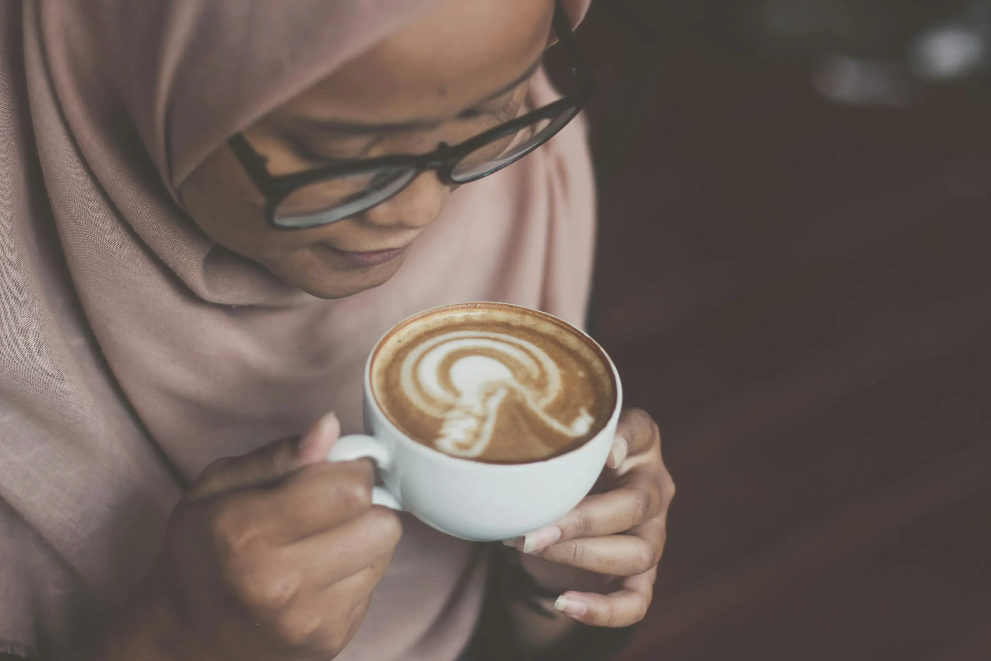 A high-angle, close-up shot shows a woman wearing glasses and a light-colored hijab looking down at a white cup of coffee she is holding. The latte art on the surface of the coffee features a light bulb design surrounded by concentric half-circles.