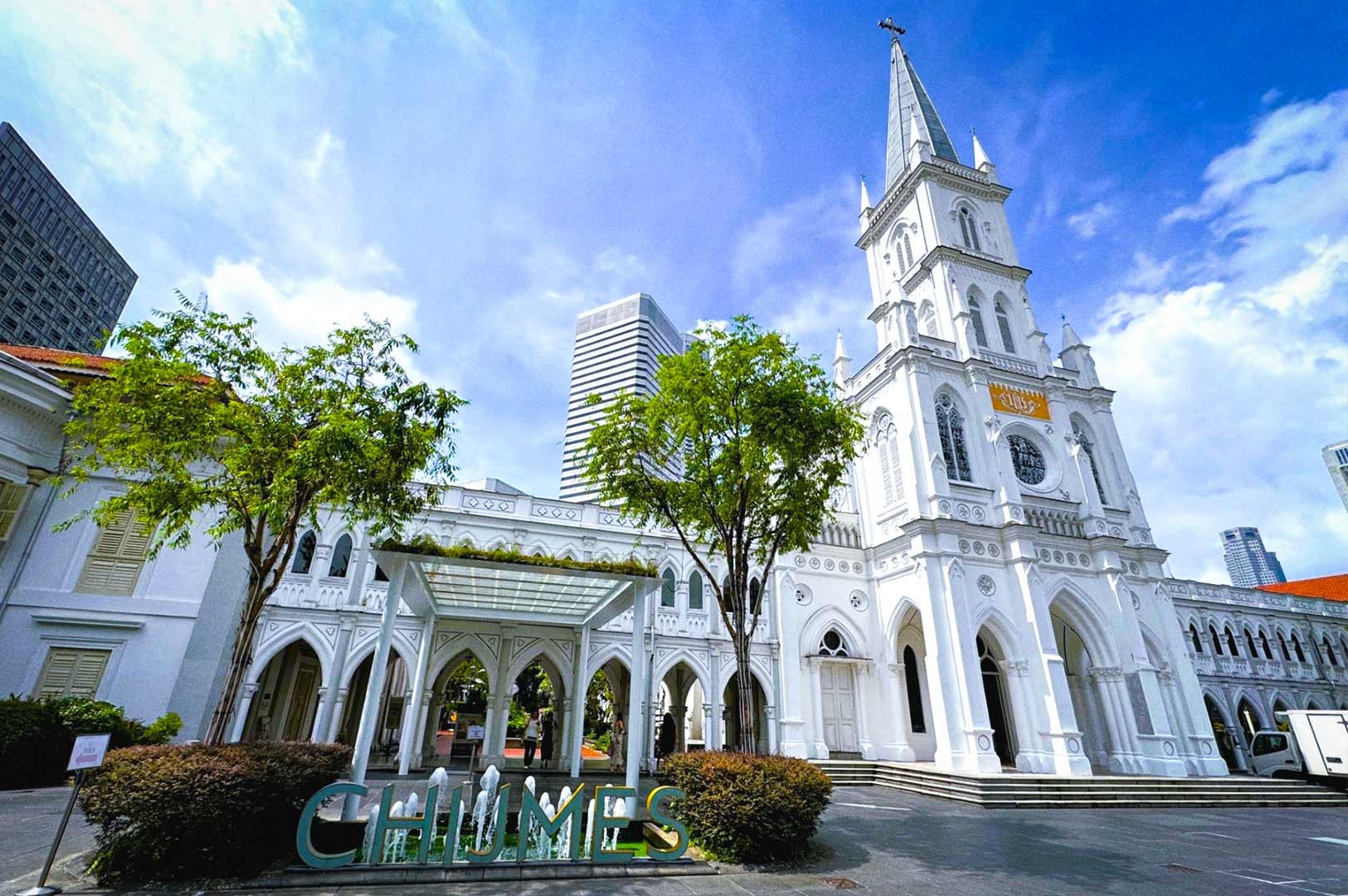 Daytime view of CHIJMES, a historic white Gothic-style complex with a tall chapel spire, arched windows, and decorative detailing. Trees and a glass canopy frame the entrance, with modern skyscrapers visible in the background under a bright blue sky.