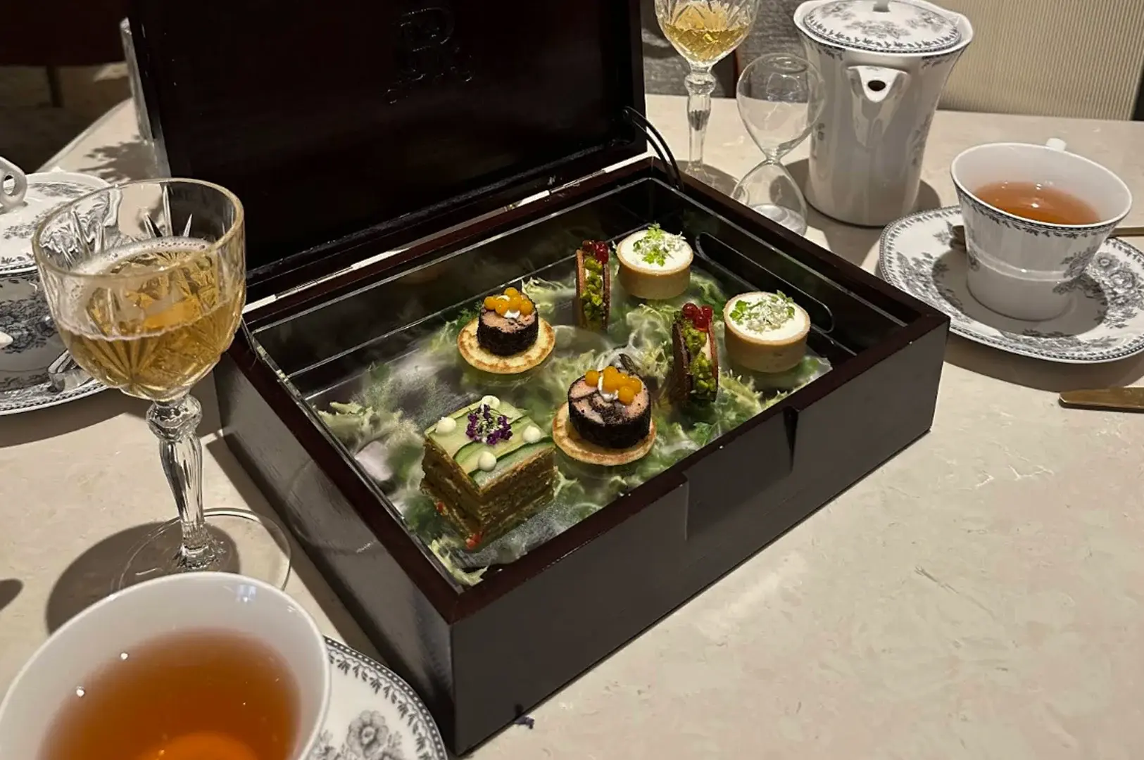 A high-angle, close-up shot displays an array of delicate afternoon tea pastries presented inside an open, dark wooden box, which rests on a light-colored marble table. Inside the box, a clear glass pane reveals a bed of green leaves subtly surrounded by a soft layer of dry ice fog, showcasing six meticulously crafted savory and sweet bites: two cylindrical rolled pastries topped with yellow caviar, two small round tarts with white cream and green herbs, a layered rectangular finger sandwich topped with sliced cucumber and purple flowers, and two small round sandwiches filled with a dark mixture. The table setting is completed with elegant, patterned porcelain teacups filled with amber tea, a crystal glass filled with a light golden beverage, a crystal hourglass timer, and a matching porcelain teapot in the background.