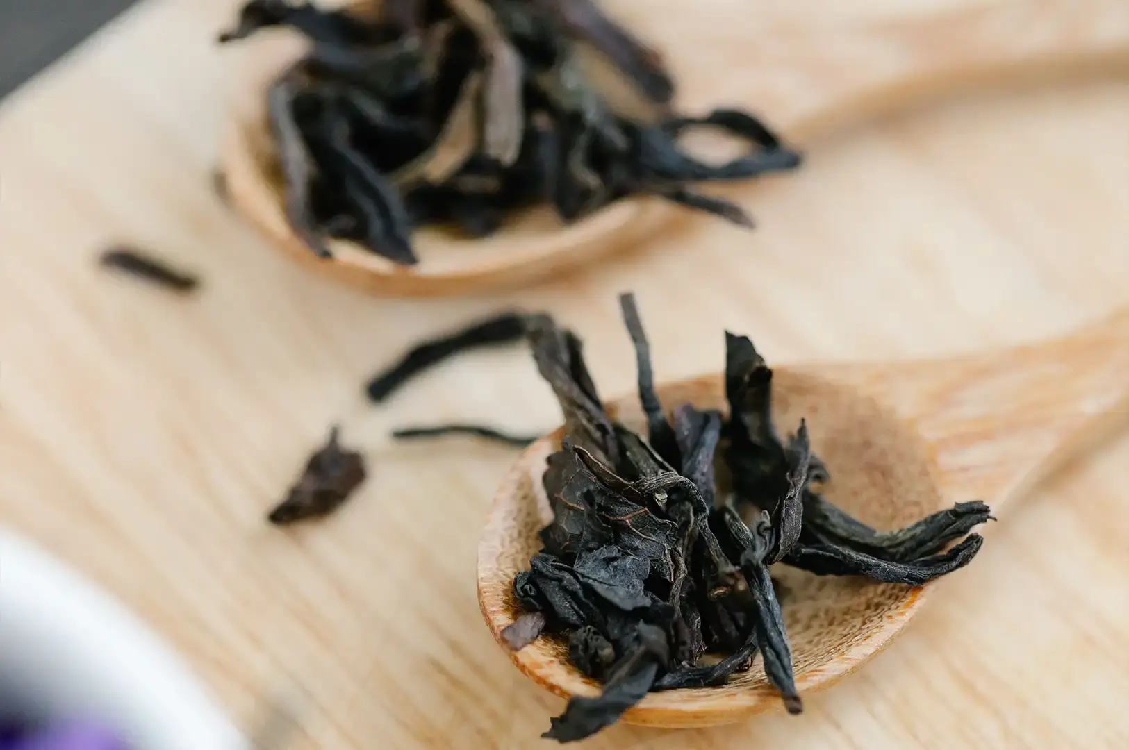 A close-up view resting on a light wooden surface shows two wooden spoons heaped with dark, loosely twisted dried tea leaves.