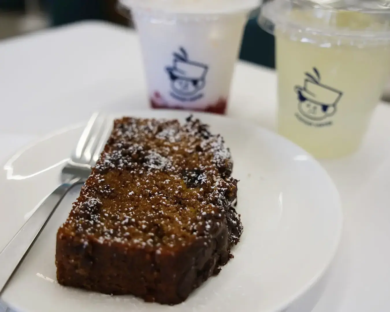 A slice of rich, chocolate chip banana bread on a white plate with a fork, accompanied by two logo-printed plastic cups of drinks in the background.