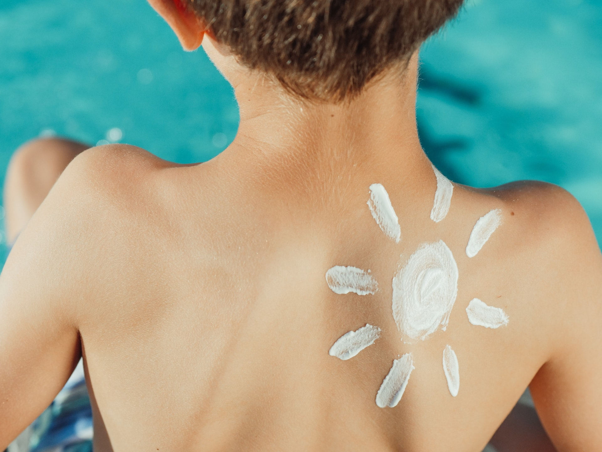 Detalhe das costas de menino de cabelos curtos, com idade aparente de 8 a 10 anos, sentado na beira de uma piscina, sem camisa, com sol de protetor solar desenhado sobre a pele, próximo ao ombro. Imagem - Pexels.com